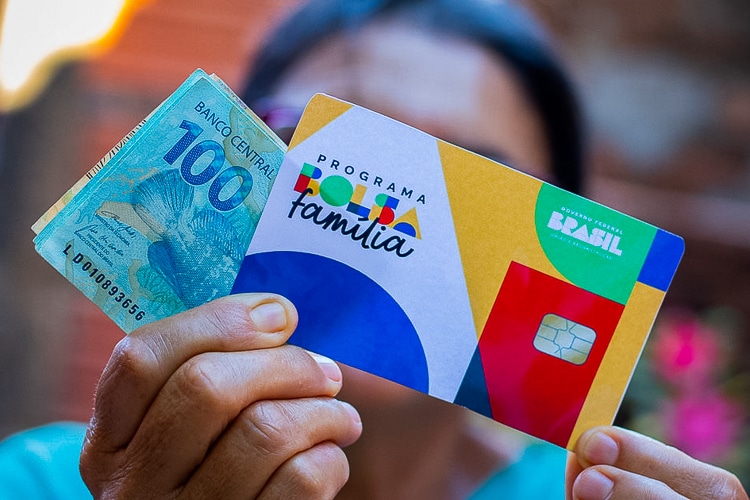 mulher segurando cartao de beneficio bolsa familia e nota de 100 e 50 reais