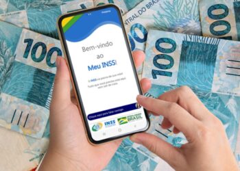 Várias notas de 100 ao lado de uma mão segurando o celular aberto no aplicativo do INSS para fazer o pedido do consignado