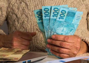 mãos segurando notas de cem reais que caem junto com o 13 salário INSS.