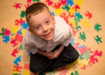 criança com autismo sorrindo para câmera e ao lado de várias peças coloridas de quebra-cabeça, que representa o transtorno
