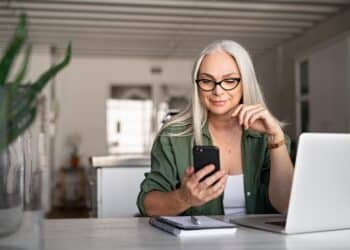 mulher de cabelos brancos sentada no escritório ao lado do computador e com celular na mão