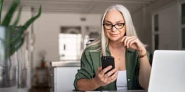 mulher de cabelos brancos sentada no escritório ao lado do computador e com celular na mão