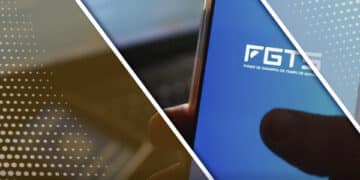 mão segurando smartphone com app do fgts aberto, ao fundo há um computador com o site do fgts aberto e sobre o teclado do computador há uma carteira de trabalho