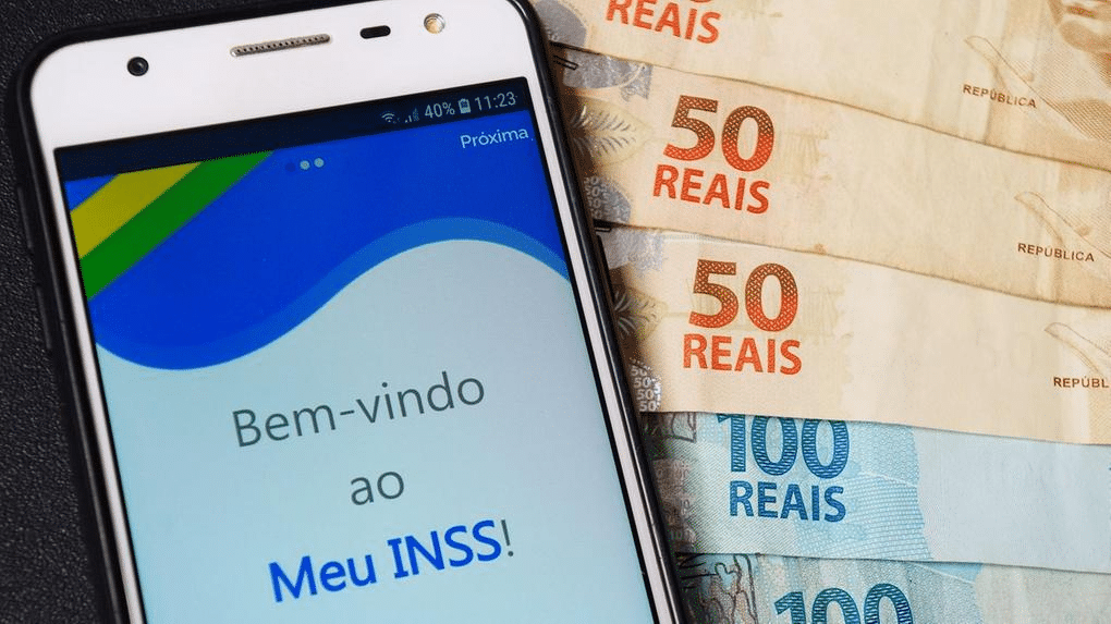 Celular aberto no app do INSS e com notas de R$100 e R$200 ao lado. 