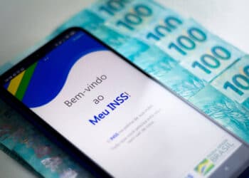 tela do celular aberta no site do INSS ao lado de várias notas de 100 reais