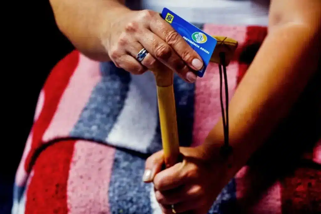 Imagem de idosa da cintura para baixo apoiada na bengala e segurando o cartão da previdência social