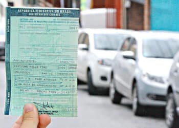 mão segurando um documento (ipva), ao fundo há uma fila de carros