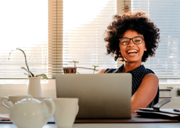 mulher negra, sorridente, com cabelo estilo blackpower, usando óculos, sentada frente a uma escrivaninha com um laptop em cima.