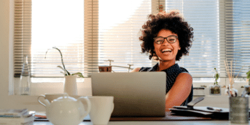 mulher negra, sorridente, com cabelo estilo blackpower, usando óculos, sentada frente a uma escrivaninha com um laptop em cima.