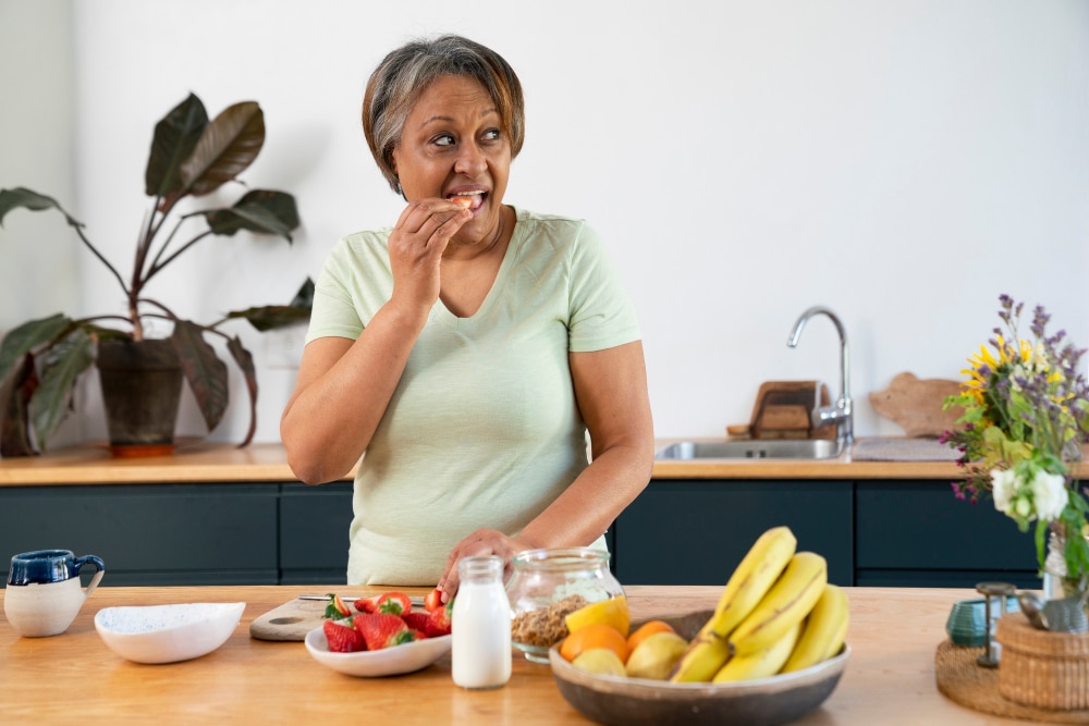 mulher negra, por volta dos 60 anos, comendo morango, à frente dela há um prato com morangos, como de leite e bandeja com bananas