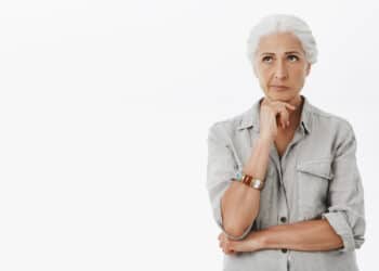 mulher aparentando ter uns 65 anos, de cabelos brancos, camisa cinza, com a mão sob o queixo, parecendo estar em dúvida