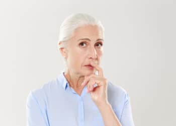 mulher com mais ou menos 65 anos, usando camisa azul, de cabelos brancos, com a mão sob o queixo, com ares de dúvida