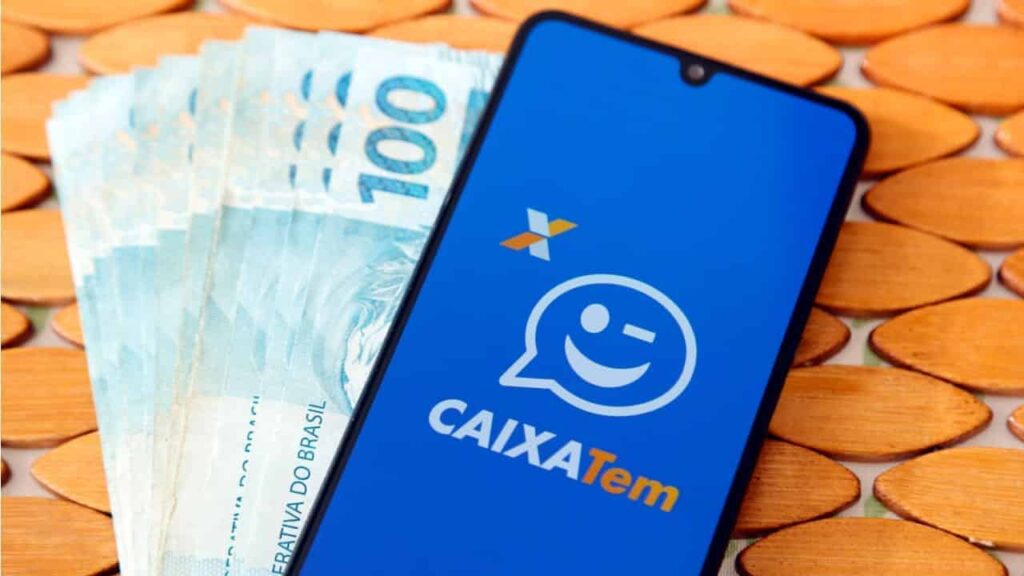 Celular aberto no Caixa Tem ao lado de notas de 100 reais para receber o PIS/PASEP 2025. (Fonte: shutterstock).
