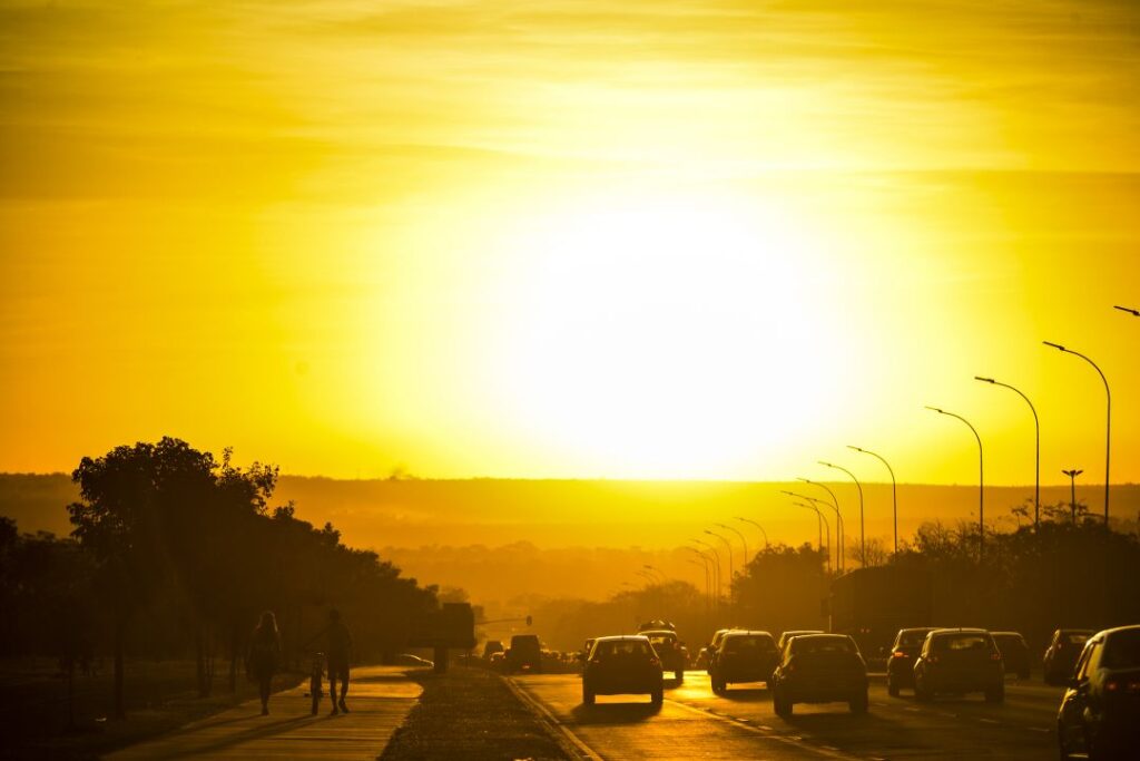 Trânsito na cidade com vários carros e pedestres no horário de verão com céu ensolarado. (Fonte: Revista Amanhã).