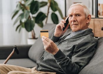 Idoso sentado no sofá e ao telefone segurando cartão de crédito para negativado. (Fonte: shutterstock).