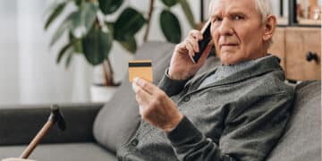 Idoso sentado no sofá e ao telefone segurando cartão de crédito para negativado. (Fonte: shutterstock).