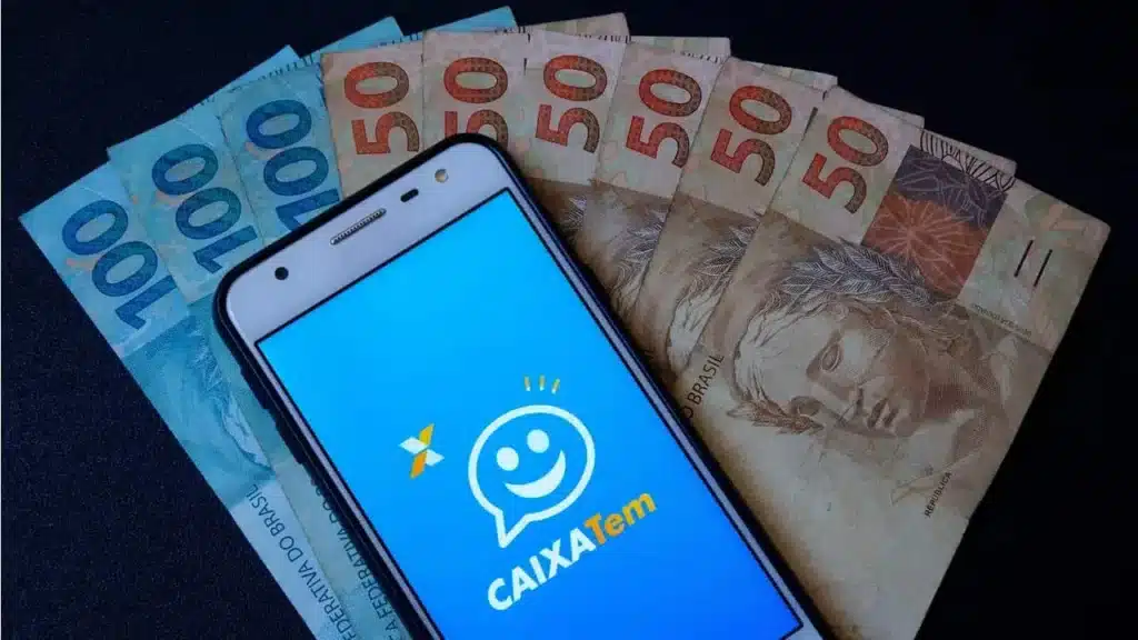 celular aberto no aplicativo caixa tem ao lado de notas de 50 e 100 reais