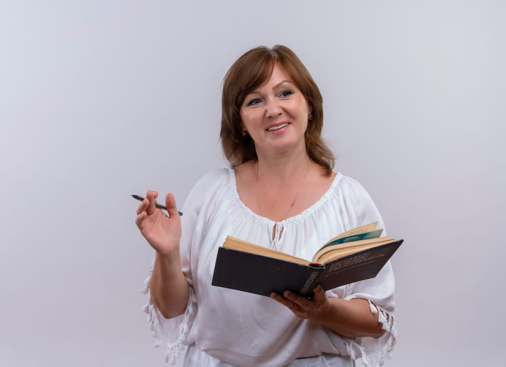 professora de meia idade, segurando um livro com uma mão e com a outra uma caneta.