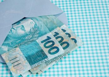 Pagamento extra do INSS em notas de 100 reais, em cima de uma mesa e dentro de uma carteira azul clara. (Fonte:  Getty Images/iStockphoto/AndreaGoldschmidt).