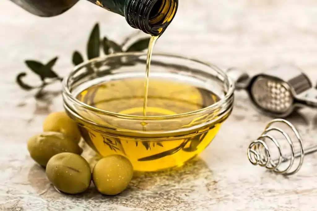 Azeite de oliva sendo despejado em um recipiente de vidro, ao lado de azeitonas. (Fonte: pixabay).