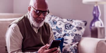 Homem mais velho sentado em um sofá bege, mexendo no celular para pedir empréstimo. (Fonte: Vivo).
