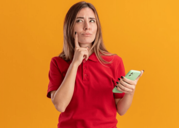 mulher loura usando tshirt vermelha, segurando celular com uma mão e a outra sobre o queixo