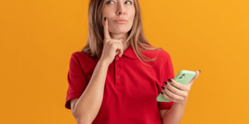 mulher loura usando tshirt vermelha, segurando celular com uma mão e a outra sobre o queixo
