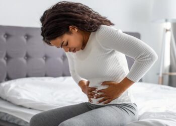 mulher negra sentada sobre um cama, com as mãos sobre o ventre e muita dor (Fonte: Freepik)