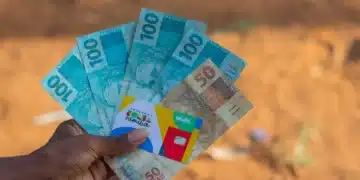 closeup em mão de pessoa negra segurando cartão do bolsa família e notas de 100 e 50 reais