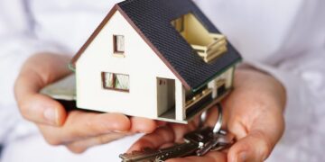 foto aproximada de uma pessoa segurando uma pequena maquete de casa numa mãe e um molho de chaves na outra