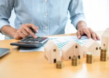 mulher fazendo contas usando calculadora, papel e caneta. na mesa há casinhas de madeira e moedas