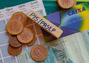 moedas, nota de 100 e cartão da caixa sobre calendário. sobre tudo há um bloco de madeira onde está escrito a caneta 'pis/pasep'