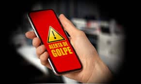 closeup de mão segurando smartphone com alerta de golpe escrito sobre fundo vermelho