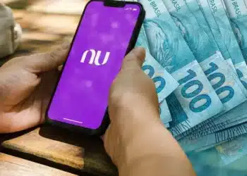 close up mão segurando smartphone com app do nubank aberta e ao fundo notas de 100