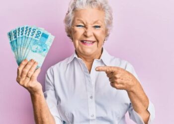 Senior grey-haired woman holding 100 brazilian real banknotes smiling happy pointing with hand and finger